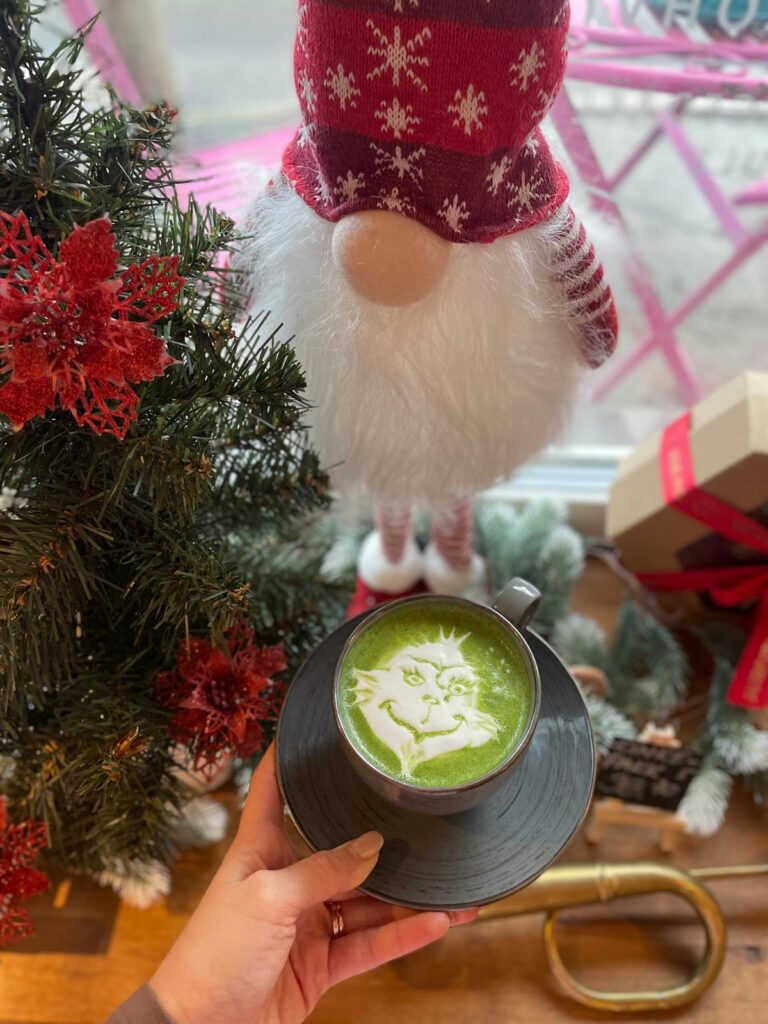 Green Grinch latte in a black cup with a Christmas gnome and tree behind